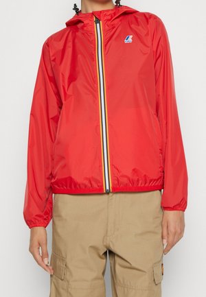 Person wearing a red hooded jacket with striped zipper and beige cargo pants, standing against a plain light background.