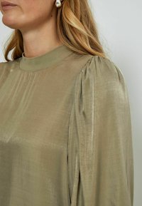 Olive green blouse with a high neck, featuring pleated detailing at the shoulders. Made of smooth, shiny fabric with a subtle texture.