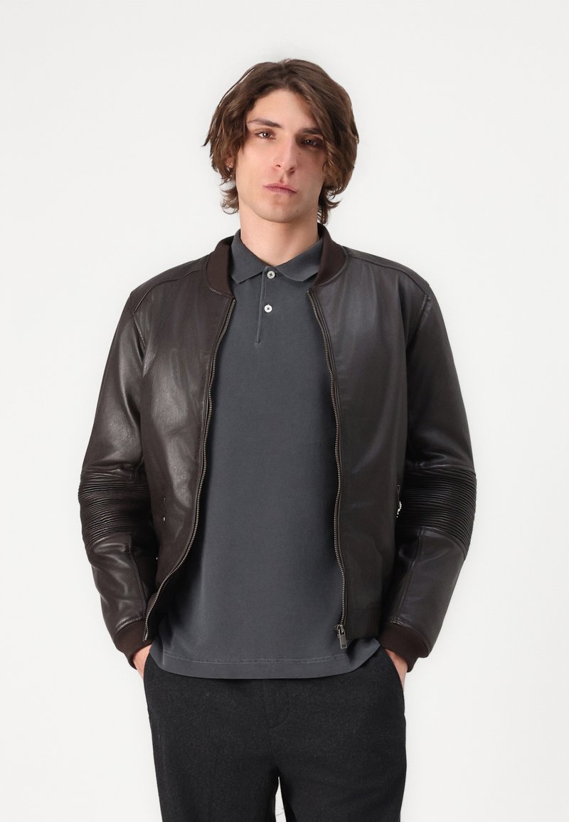 Bruine leren bomberjack met ribbels op de manchetten en schouders, voorzien van een ritssluiting aan de voorkant. Eronder is een grijze polo zichtbaar.