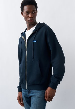 Man draagt een marineblauwe hoodie met rits en een klein "Hugo" label, een wit T-shirt en een blauwe spijkerbroek, kijkt naar links tegen een effen achtergrond.