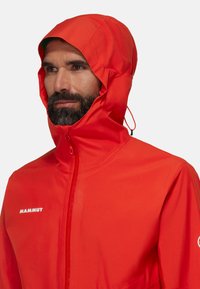 Giacca impermeabile rossa con colletto alto e cappuccio regolabile. Presenta il logo "Mammut" in bianco sul lato sinistro del petto e una texture liscia.