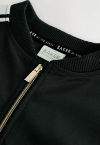 Čierna zipsová bunda s rebrovaným golierom. Obsahuje zlaté kovania a označenie značky s bielym písmom. Hladká tkanina, minimalistický dizajn.