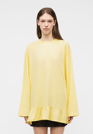 Kobieta o długich brązowych włosach, ubrana w luźną, długorękawową żółtą bluzkę z falbanką na dole oraz krótką czarną spódnicę, zwrócona przodem.