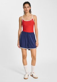 Rode tanktop met witte details, gecombineerd met marineblauwe shorts met elastische tailleband. Model draagt witte crew-sokken en casual schoenen.