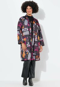 Kimono negro con estampados coloridos, que presenta patrones geométricos y texto en naranja y rosa. Llevado sobre pantalones brillantes negros.