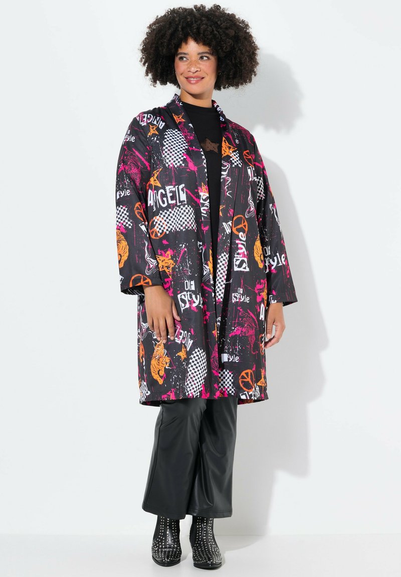Kimono negro con estampados coloridos, que presenta patrones geométricos y texto en naranja y rosa. Llevado sobre pantalones brillantes negros.