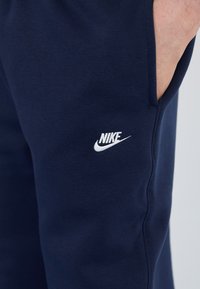 Tummansinisiä puuvillaisia collegehousuja, joissa on sivutasku ja valkoinen Nike-logo reidessä. Sileä pinta ja kapeneva malli nilkassa.