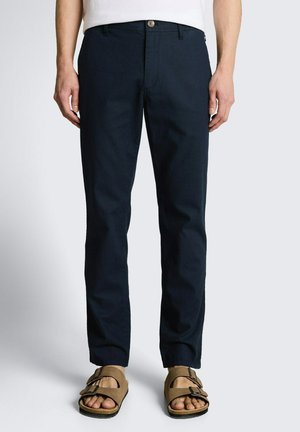 Mężczyzna w granatowych spodniach o prostym kroju i jasnobrązowych sandałach z podwójnym paskiem, stojący na jednolitym białym tle.