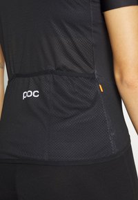 Acercamiento a una persona que lleva puesta una camiseta de ciclismo negra texturizada con tres bolsillos traseros y un pequeño tirador de cremallera naranja.