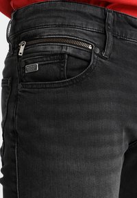 Pantalones vaqueros de mezclilla negra con un bolsillo con cremallera, dos botones delanteros y una textura suave. Etiqueta de la marca visible encima del bolsillo.