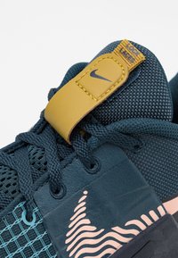 Mörkblå sportskor med meshtyg, texturerade detaljer och ett gult snabblås. Anmärkningsmönster i rosa v ågor på sidan.