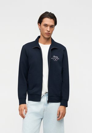 Man draagt een marineblauw jasje met rits met het "Tommy Hilfiger" logo, een wit t-shirt en lichtblauwe spijkerbroek, staand tegen een effen lichte achtergrond.