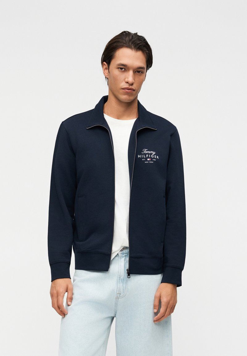 Homme portant une veste zippée bleu marine avec le logo "Tommy Hilfiger", un t-shirt blanc et un jean bleu clair, debout devant un fond uni clair.