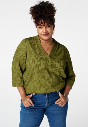 Krullend persoon met een olijfgroen top met textuur en blauwe spijkerbroek, staand met handen in de zakken tegen een witte achtergrond.