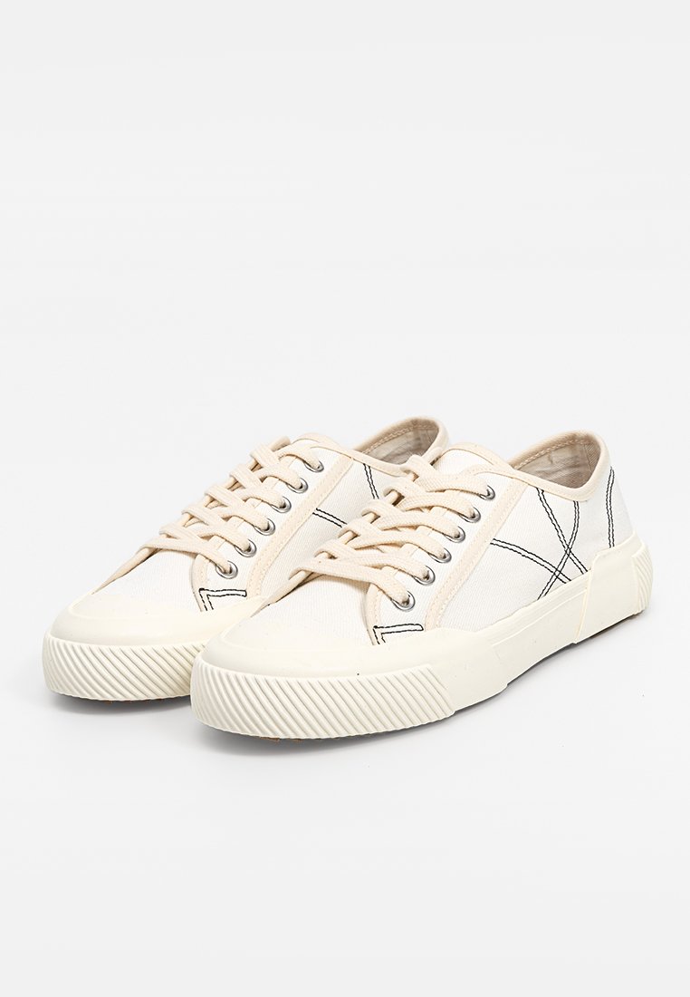 Sneakers in tela beige con cuciture nere, punta rotonda, suola in gomma e lacci piatti. Presentano dettagli testurizzati e una suola a contrasto.