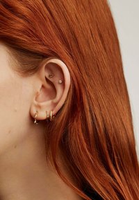 Pendientes de oro con formas variadas: un aro texturizado, una banda sencilla y un pendiente con una pequeña gema, en contraste con el cabello castaño rojizo.