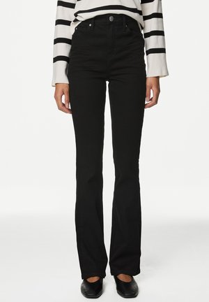 Personne portant un pantalon noir taille haute évasé, une chemise blanche à manches longues avec des rayures noires horizontales et des chaussures noires à bout carré.