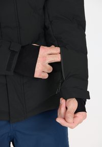 Schwarze isolierte Jacke mit einer Reißverschlusstasche am Ärmel. Der Handgelenkabschluss zeigt eine Stoffüberlagerung und eine enge Passform. Der Hintergrund ist neutral.