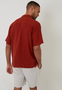 Kortærmet, rustfarvet skjorte lavet af hør, med en klassisk krave og en afslappet pasform. Båret sammen med lyse shorts.
