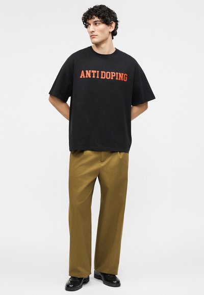 Homme aux cheveux bouclés portant un t-shirt noir "ANTI DOPING", un pantalon large couleur moutarde et des chaussures noires, sur un fond uni.