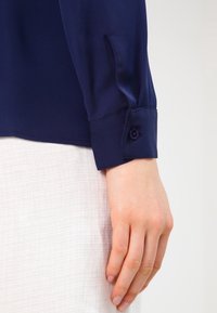 Blouse en satin bleu marine avec des poignets à bouton, présentant une texture lisse et une coupe ajustée, tenue par une main sur un fond clair.