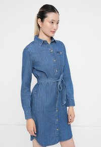 Ung kvinne med bundet hår har på seg en belted, knappet denimkjole med lange ermer og brystlomme mot en hvit bakgrunn.