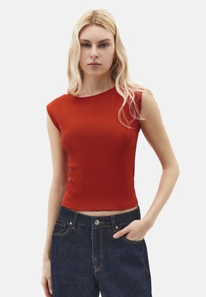 Kvinde med langt blond hår iført en ærmeløs, tætsiddende rød top og mørkeblå jeans, stående med den ene hånd i lommen mod en ensfarvet baggrund.