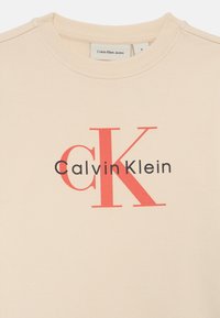 Sudadera beige con un gran logotipo rojo de "CK" y texto en negro que dice "Calvin Klein". Tejido suave y liso con un cuello redondo acanalado.