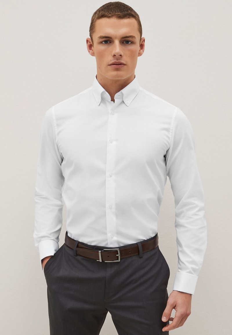 Biela košeľa s dlhým rukávom a golierom na gombíky, vyrobená z hladkej tkaniny. Má slim fit strih, nosená s tmavosivými nohavicami a hnedým opaskom.