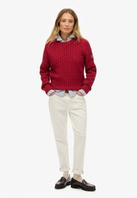 Rode gebreide trui met kabelpatroon, blauwe en witte gestreepte shirt eronder, crèmekleurige broek en donkere loafers met opgerolde mouwen.