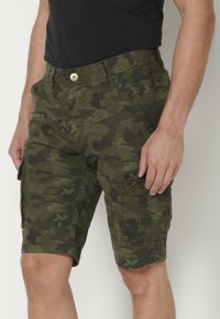 Mies, jolla on päällään vihreät camouflage-housut ja musta t-paita, seisoo kädet rentoina sivuilla tasaisessa taustassa.
