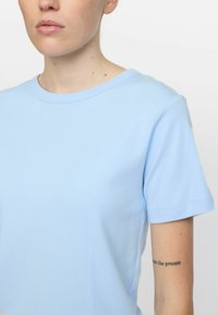 Svijetloplava pamučna majica s okruglim izrezom, kratkim rukavima i glatkom teksturom, koja sadrži suptilne šavove i prilagođeni kroj.