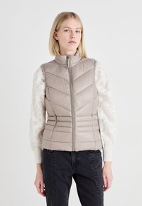 Gilet matelassé beige clair avec un col haut, une fermeture éclair et des coutures en forme de chevron. Accentué par des manches en tricot crème texturé.