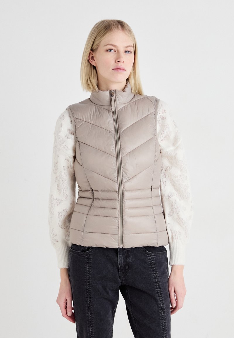 Gilet matelassé beige clair avec un col haut, une fermeture éclair et des coutures en forme de chevron. Accentué par des manches en tricot crème texturé.