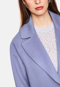 Hellpurpurner Wollmantel mit Schalkragen, strukturiertem Nähten, über einem hellen Strickpullover mit Spitzenmustern getragen.