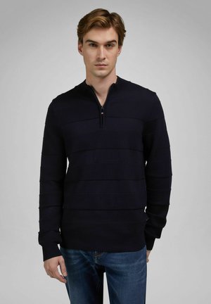 Junger Mann mit hellbraunen Haaren, der einen marineblauen Langarm-Sweater mit Reißverschluss am Hals und eine blaue Jeans vor einem schlichten grauen Hintergrund trägt.