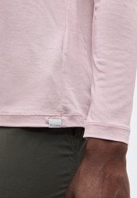 Κοντινό πλάνο του χεριού ενός ατόμου που φοράει μακρυμάνικο μπλουζάκι ανοιχτό ροζ και σκούρο παντελόνι, δείχνοντας μια μικρή ετικέτα με το λογότυπο της Houdini στο τελείωμα.