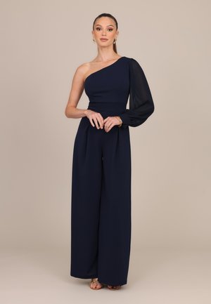 Femme portant une combinaison bleu marine à une épaule avec une manche longue transparente, des boucles d'oreilles dorées, des bracelets et des talons ouverts, debout sur un fond uni.