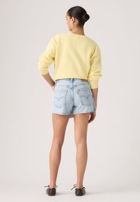 Γυναίκα με τα μαλλιά της σε κότσο, φοράει ανοιχτό κίτρινο πουλόβερ, ανοιχτά μπλε σορτς από τζιν και σκούρα καφέ δερμάτινα παπούτσια με κορδόνια, στέκεται μπροστά σε απλό φόντο.