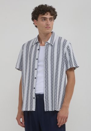 Un homme aux cheveux bouclés portant un pantalon bleu marine, un t-shirt sans manches blanc et une chemise blanche à manches courtes avec des rayures verticales géométriques noires.