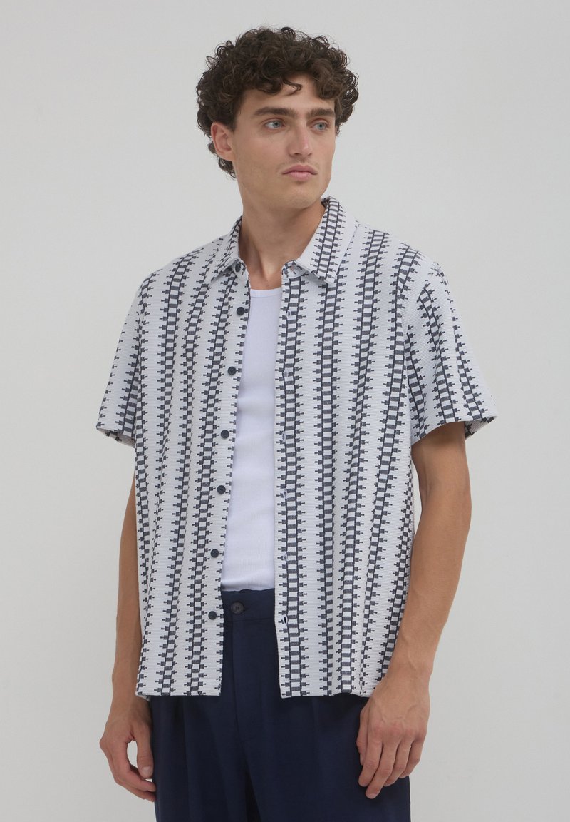 Un homme aux cheveux bouclés portant un pantalon bleu marine, un t-shirt sans manches blanc et une chemise blanche à manches courtes avec des rayures verticales géométriques noires.
