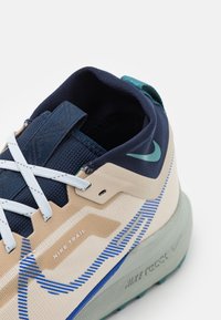 Chaussure de trail Nike avec une tige beige et marine, en mesh texturé, des accents bleus et une languette de talon verte.