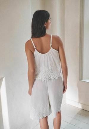 Femme aux cheveux foncés portant un haut blanc sans manches et un short blanc assorti avec des détails en dentelle, debout à l'intérieur, dos à la caméra.