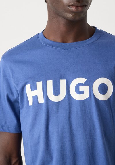 Синя памучна тениска с изразен бял логотип "HUGO" на предната част. Тканината изглежда мека, с кръгло деколте.