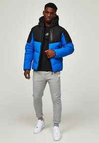 Puffermantel mit einem schwarz-blauen Farbblockdesign, mit Kapuze und Reißverschluss. Graue Jogginghose und weiße Sneaker vervollständigen das Outfit.