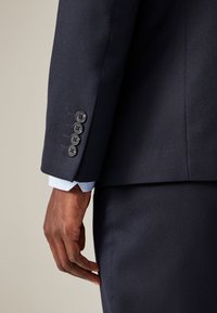 Meri sinine tekstuuriga bleiseri varrukal on neli musta nööpi ja sealt paistab helesinine mustriga mansett.