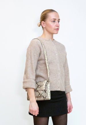 Frau trägt einen beigen knöpfbaren Pullover und einen schwarzen Rock, hält eine Schultertasche mit Schlangenledermuster und steht vor weißem Hintergrund.