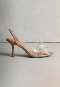 Naakte slingback hoge hak schoen met transparante bandjes en een kralenbloemversiering op de wreef. Voorzien van een spits toelopende neus en een naaldhak.
