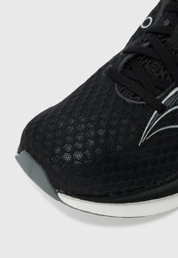 Chaussure de sport noire avec une tige en mesh, bout arrondi, lacets noirs et semelle extérieure grise. Accent argenté notable sur le côté pour un détail de design supplémentaire.