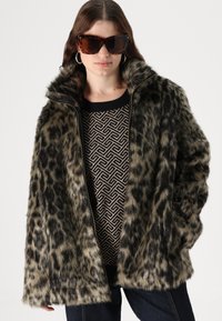 Jakna iz umetnega krzna z leopardjim vzorcem in visokim ovratnikom, z zadrgo in sproščenim fitom, nosena preko vzorčastega puloverja. Rjave sončne očala.